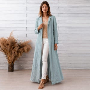 Puede incluir: Un abrigo largo de color azul claro con mangas largas y frente abierto, usado sobre una camiseta sin mangas beige y pantalones blancos ajustados. El abrigo presenta una tela texturizada, un cuello recto y un dobladillo largo hasta el suelo.