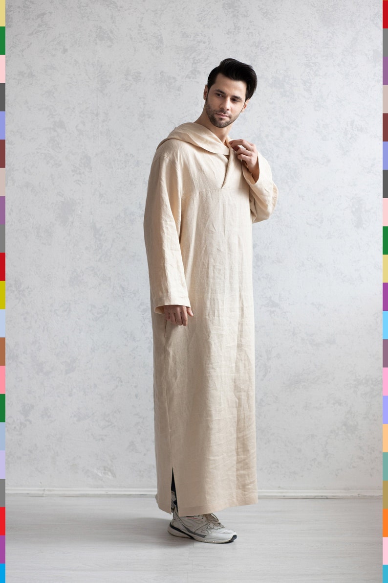 May include: A beige linen robe with a hood and a long, flowing silhouette. The robe has a V-neck and a side slit. The person is wearing white sneakers.