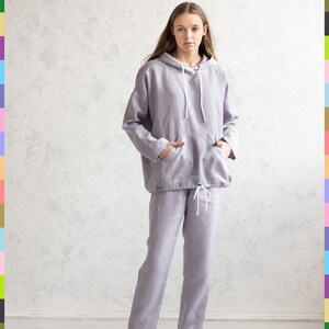 K&ouml;nnte beinhalten: Ein helllila Kapuzenpullover und eine passende Hose aus Leinen. Der Kapuzenpullover hat eine Kapuze mit Kordelzug und eine Vordertasche. Die Hose hat einen Kordelzug in der Taille. Die Person tr&auml;gt wei&szlig;e Turnschuhe. Das Outfit ist l&auml;ssig und bequem.