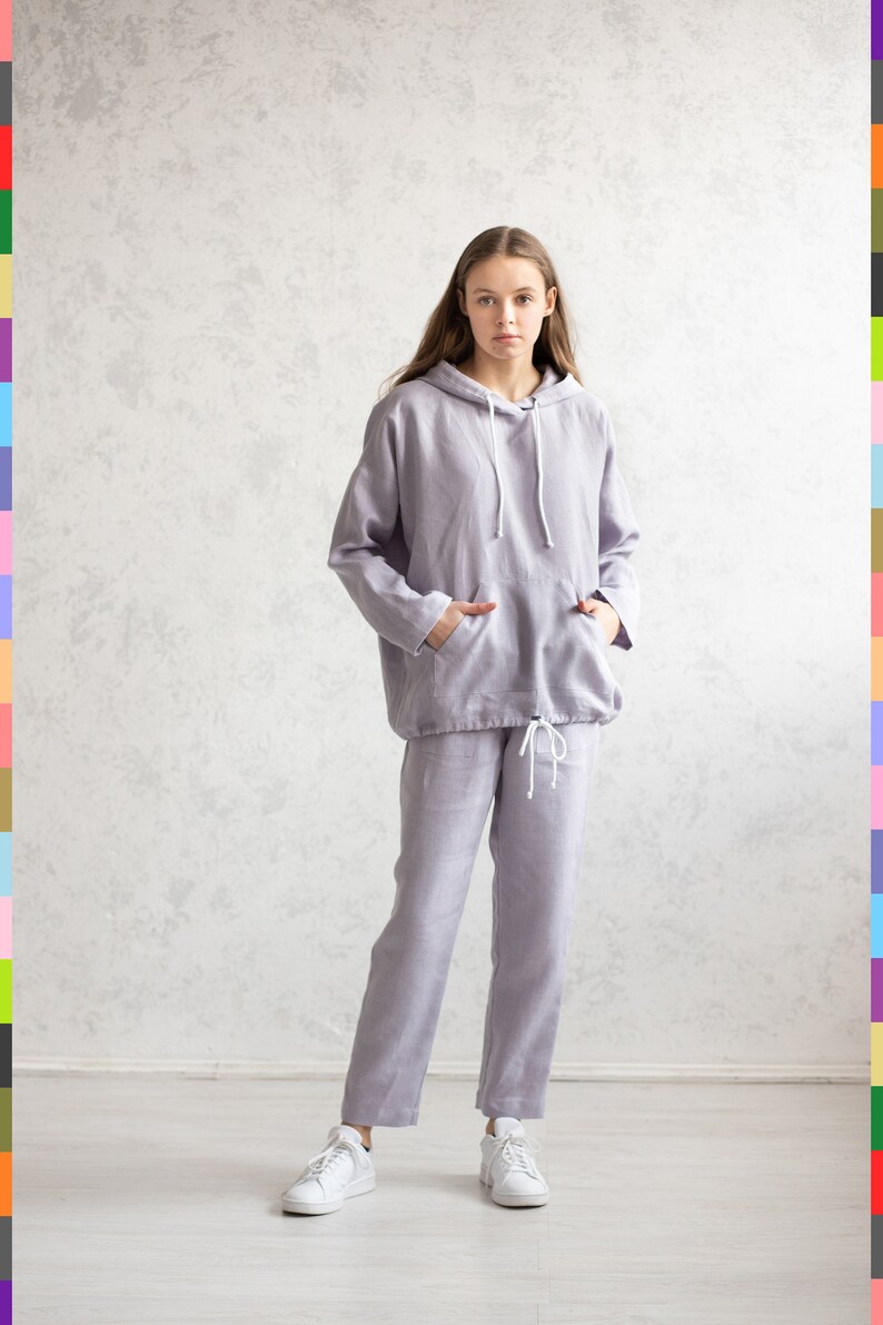 K&ouml;nnte beinhalten: Ein helllila Kapuzenpullover und eine passende Hose aus Leinen. Der Kapuzenpullover hat eine Kapuze mit Kordelzug und eine Vordertasche. Die Hose hat einen Kordelzug in der Taille. Die Person tr&auml;gt wei&szlig;e Turnschuhe. Das Outfit ist l&auml;ssig und bequem.