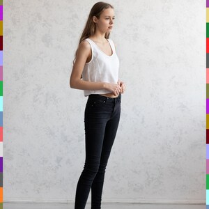 May include: A woman wearing a white sleeveless crop top and black skinny jeans. She is also wearing white sneakers. The background is a light gray wall and a light gray floor.