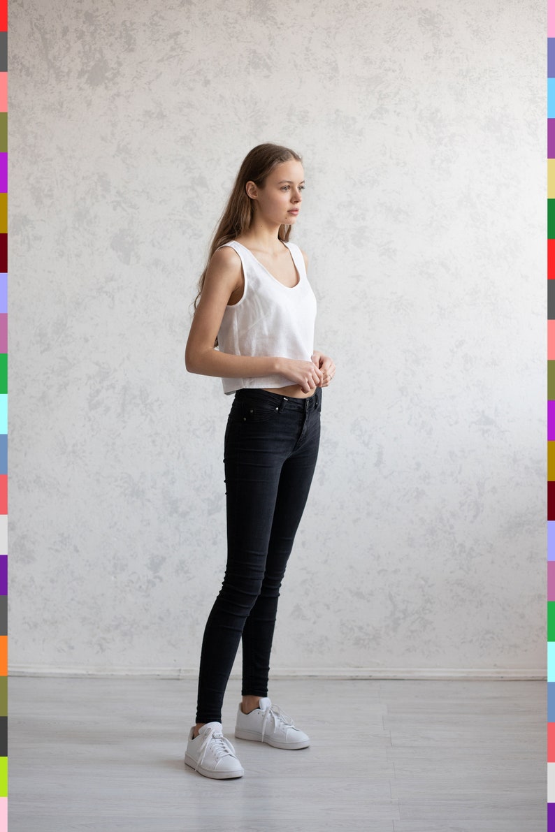 May include: A woman wearing a white sleeveless crop top and black skinny jeans. She is also wearing white sneakers. The background is a light gray wall and a light gray floor.