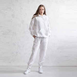 May include: A white linen hoodie and matching pants set. The hoodie has a front pocket and drawstring, while the pants have an elastic waistband and cuffs. The outfit is paired with white sneakers, creating a casual, comfortable look.