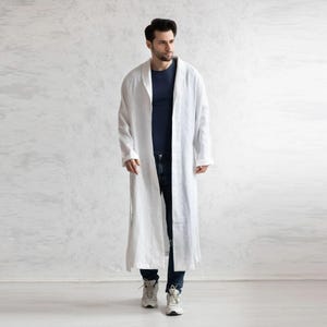 May include: A long, white linen robe with a shawl collar. The robe is floor-length and has long sleeves. The person is wearing a navy blue shirt and dark blue jeans with white sneakers. The background is a plain, textured white wall.