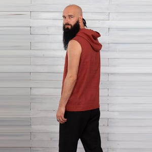May include: A sleeveless, hooded top in a reddish-brown hue. The garment has a relaxed fit and a hood. The person in the image is wearing black pants. The background is a white wooden wall.