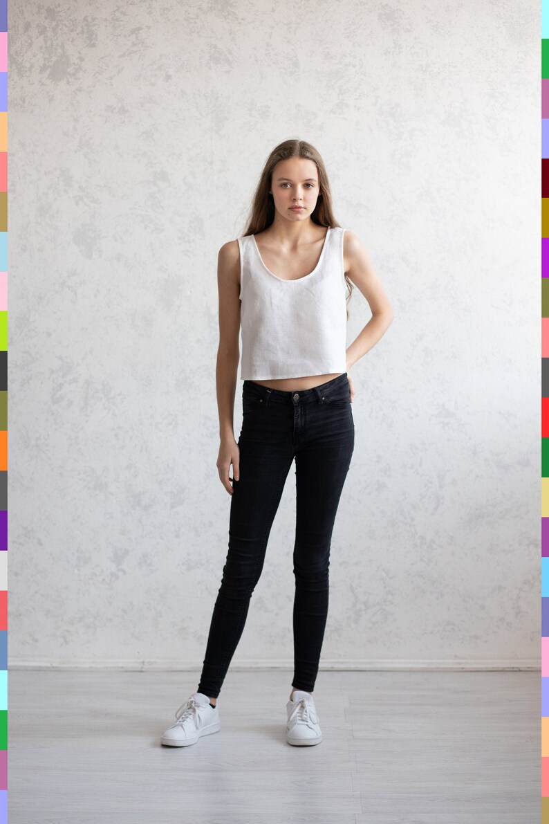 May include: A woman wearing a white sleeveless crop top and black skinny jeans. She is standing in front of a white wall and is wearing white sneakers.