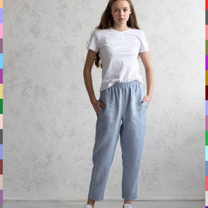 May include: A woman wearing a white t-shirt and light blue linen pants with pockets. She is also wearing white sneakers. The background is a light gray textured wall.