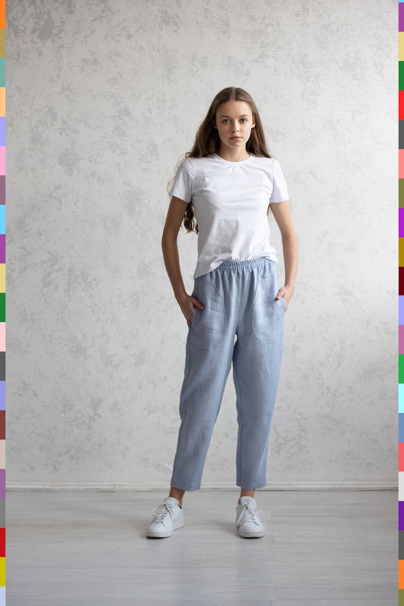 May include: A woman wearing a white t-shirt and light blue linen pants with pockets. She is also wearing white sneakers. The background is a light gray textured wall.