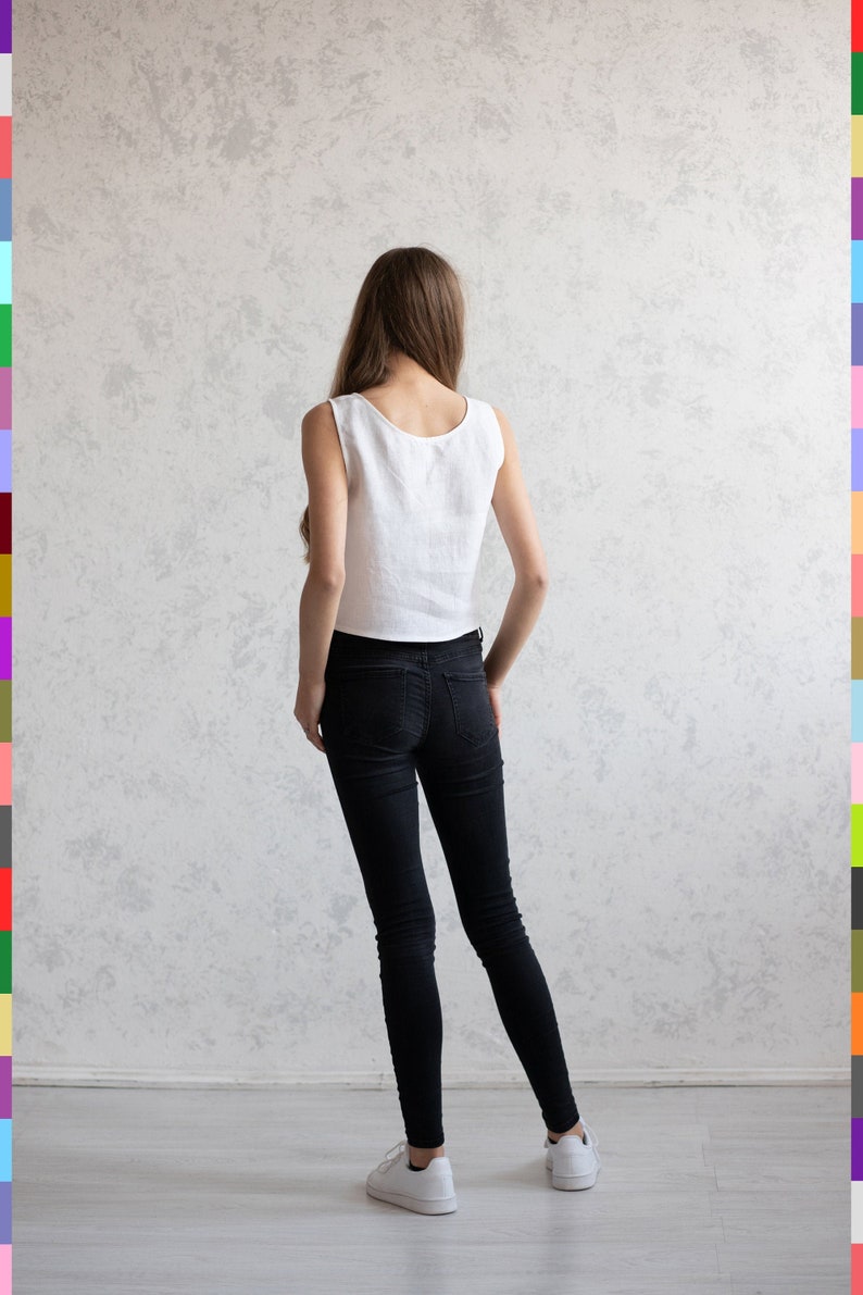 May include: A sleeveless white linen top and black skinny jeans are worn with white sneakers. The model is standing with their back to the camera against a textured gray wall. The outfit is simple and casual.