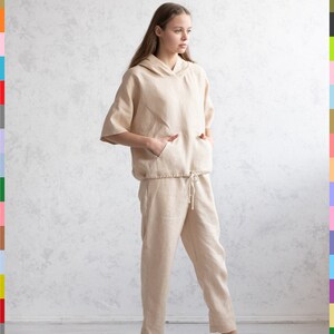 May include: A beige linen hooded top and matching pants set. The top has short sleeves and a front pocket. The pants have a drawstring waist. The outfit is paired with white sneakers. The model is standing in front of a textured wall.