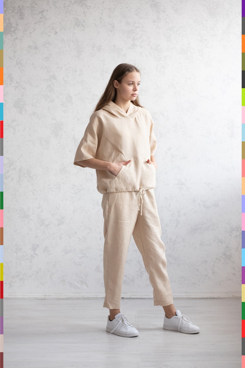 May include: A beige linen hooded top and matching pants set. The top has short sleeves and a front pocket. The pants have a drawstring waist. The outfit is paired with white sneakers. The model is standing in front of a textured wall.