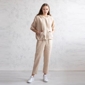 May include: A beige linen hoodie with short sleeves and a drawstring waist paired with matching linen pants. The outfit features a relaxed fit and pockets on the hoodie. The model is wearing white sneakers. This casual outfit is suitable for everyday wear.