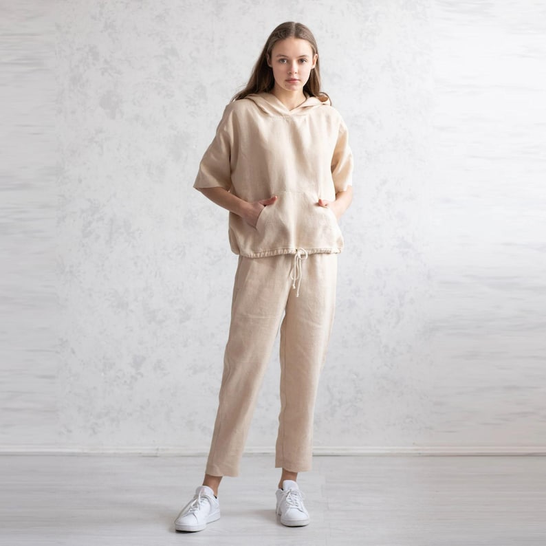 May include: A beige linen hoodie with short sleeves and a drawstring waist paired with matching linen pants. The outfit features a relaxed fit and pockets on the hoodie. The model is wearing white sneakers. This casual outfit is suitable for everyday wear.