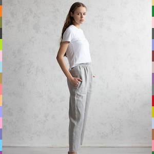 May include: A person wearing a white t-shirt and light gray linen pants. The pants have an elastic waistband and pockets. White sneakers complete the outfit. The background is a textured, light gray wall.