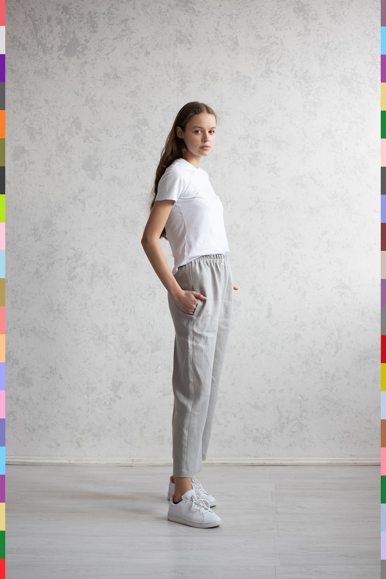 May include: A person wearing a white t-shirt and light gray linen pants. The pants have an elastic waistband and pockets. White sneakers complete the outfit. The background is a textured, light gray wall.