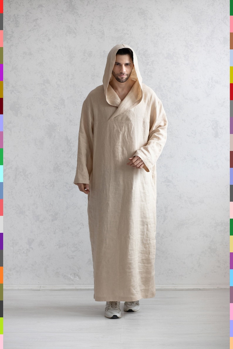 May include: A beige linen robe with a hood. The robe has a long, flowing design and is worn by a person standing in front of a white wall.