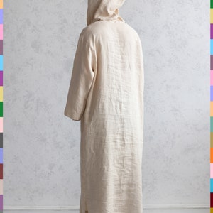 May include: A long, cream-colored linen robe with a hood. The robe is worn by a person standing on a white floor.