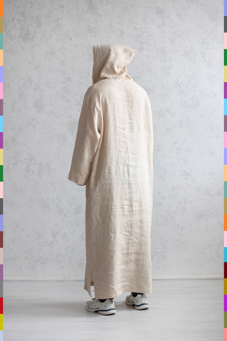 May include: A long, cream-colored linen robe with a hood. The robe is worn by a person standing on a white floor.