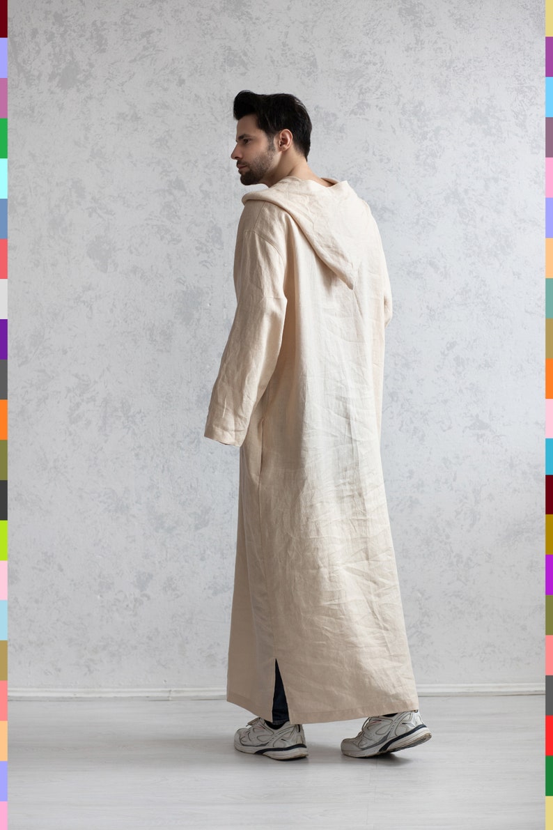 May include: A long, cream-colored hooded robe with a long sleeve and a side slit. The robe is made of a soft, lightweight fabric. The person wearing the robe is standing on a light wood floor.