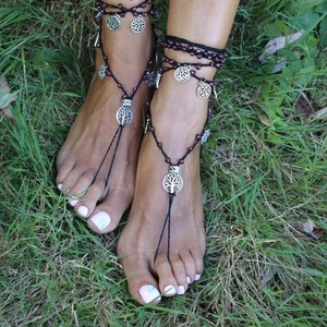 Puede incluir: Un par de sandalias descalzas negras con dijes de plata de &aacute;rbol de la vida. Las sandalias est&aacute;n hechas con cord&oacute;n negro y peque&ntilde;as cuentas moradas. Las sandalias se usan en los pies de una persona en un &aacute;rea cubierta de hierba.