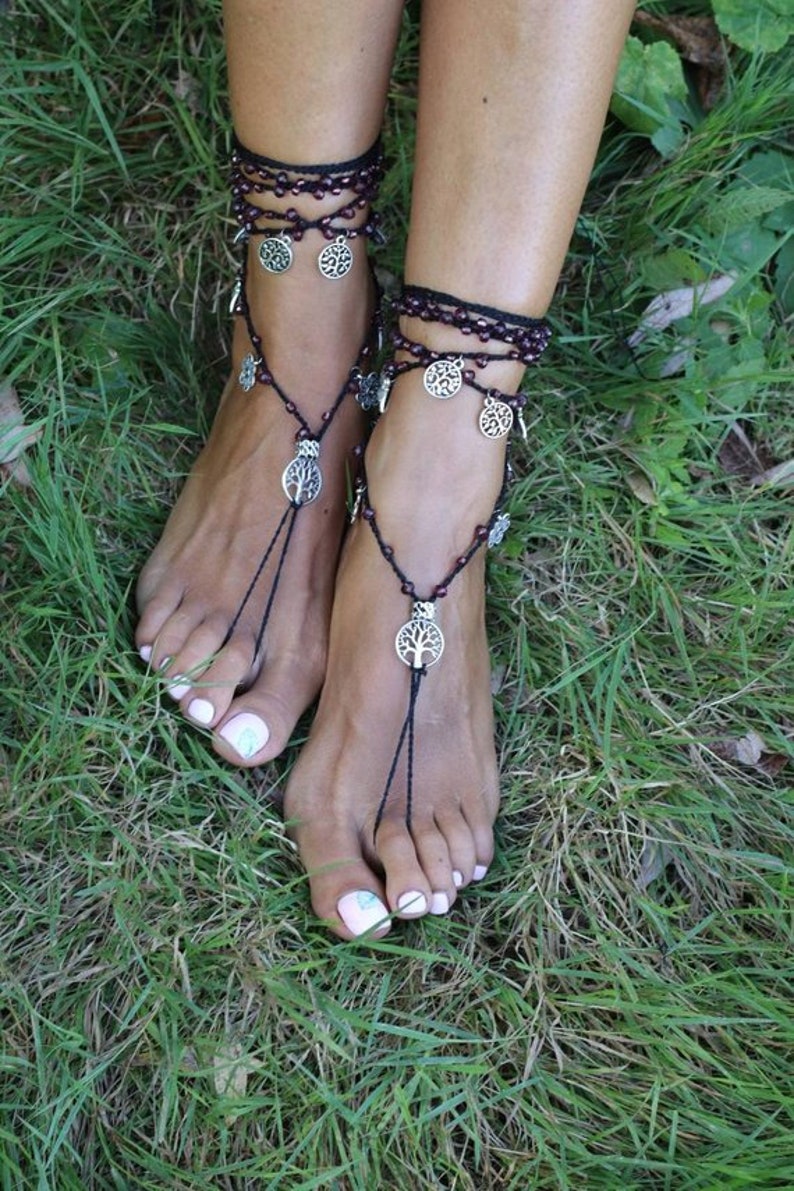 Puede incluir: Un par de sandalias descalzas negras con dijes de plata de &aacute;rbol de la vida. Las sandalias est&aacute;n hechas con cord&oacute;n negro y peque&ntilde;as cuentas moradas. Las sandalias se usan en los pies de una persona en un &aacute;rea cubierta de hierba.