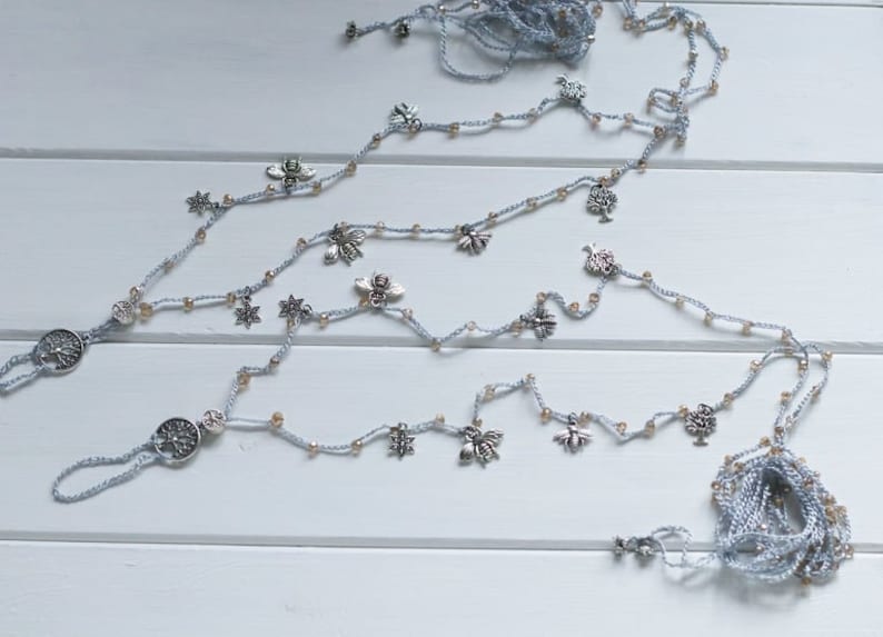 Puede incluir: Un par de joyas de pie de ganchillo con abalorios plateados y dorados. Los abalorios incluyen &aacute;rboles, estrellas y mariposas. Las joyas est&aacute;n sobre una superficie de madera blanca.