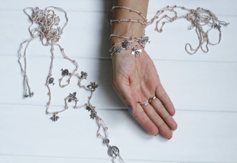 Puede incluir: Un delicado collar de cuentas de plata y oro con m&uacute;ltiples hilos y peque&ntilde;os amuletos, incluyendo p&aacute;jaros, abejas y un &aacute;rbol de la vida. El collar se coloca en capas en una mu&ntilde;eca, creando un efecto de pulsera.