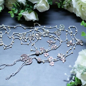 Puede incluir: Un par de joyas de pie de crochet blanco con abalorios plateados, incluyendo el &aacute;rbol de la vida, abejas y flores. Las joyas est&aacute;n dispuestas sobre una superficie gris con flores blancas en el fondo.