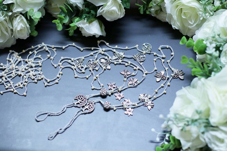 Puede incluir: Un par de joyas de pie de crochet blanco con abalorios plateados, incluyendo el &aacute;rbol de la vida, abejas y flores. Las joyas est&aacute;n dispuestas sobre una superficie gris con flores blancas en el fondo.