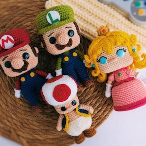 Puede incluir: Cuatro muñecos de ganchillo que representan personajes de videojuegos. Uno lleva una gorra roja con una M, otro una gorra verde con una L, otro es una figura con forma de seta con puntos rojos y otro tiene pelo amarillo y un vestido rosa.