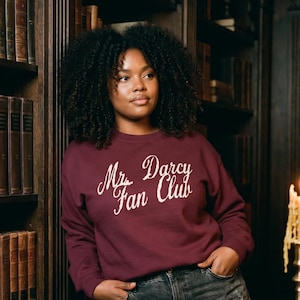 May include: A maroon long-sleeve crewneck sweatshirt featuring the white text "Mr. Darcy Fan Club" in a cursive script font across the chest, paired with dark gray distressed denim jeans and light-colored ribbed knit socks.