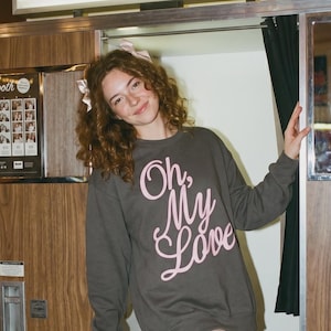 May include: A dark gray crewneck sweatshirt featuring the text Oh, My Love in light pink script lettering. The garment is worn with a white lace garter on the thigh and white platform heels with a buckled ankle strap and a block heel.