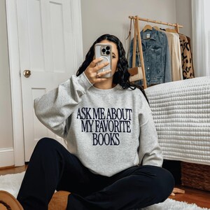May include: A light gray crewneck sweatshirt featuring the black text "ASK ME ABOUT MY FAVORITE BOOKS" printed on the chest, paired with solid black trousers and tan suede slip-on shoes with a visible patterned trim along the bottom edge.