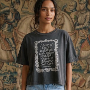 May include: A charcoal gray short-sleeve t-shirt featuring a white rectangular ornate frame graphic containing the text: I dream of Castles and Magic and Dragons and Epic. Once in a Lifetime Love Stories Breatly & Co. The shirt has a crew neckline.