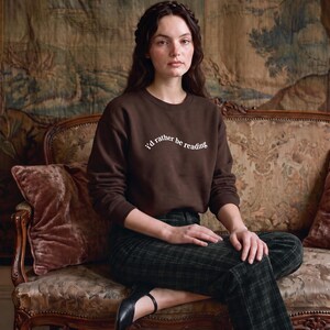 May include: A dark brown crewneck sweatshirt featuring the white text "I'd rather be reading" printed in a slight arch across the chest. The garment has long sleeves with ribbed cuffs and a ribbed neckline, constructed from a solid-colored fabric.