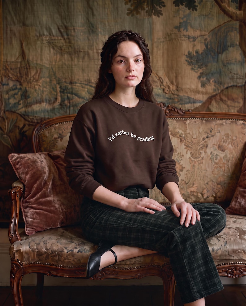 May include: A dark brown crewneck sweatshirt featuring the white text "I'd rather be reading" printed in a slight arch across the chest. The garment has long sleeves with ribbed cuffs and a ribbed neckline, constructed from a solid-colored fabric.