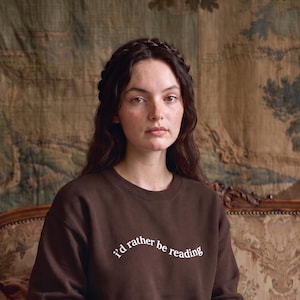 May include: A dark brown crewneck sweatshirt featuring the white text "i'd rather be reading" printed in a curved, lowercase font across the chest. The garment has long sleeves and a ribbed neckline, providing a simple and functional design.