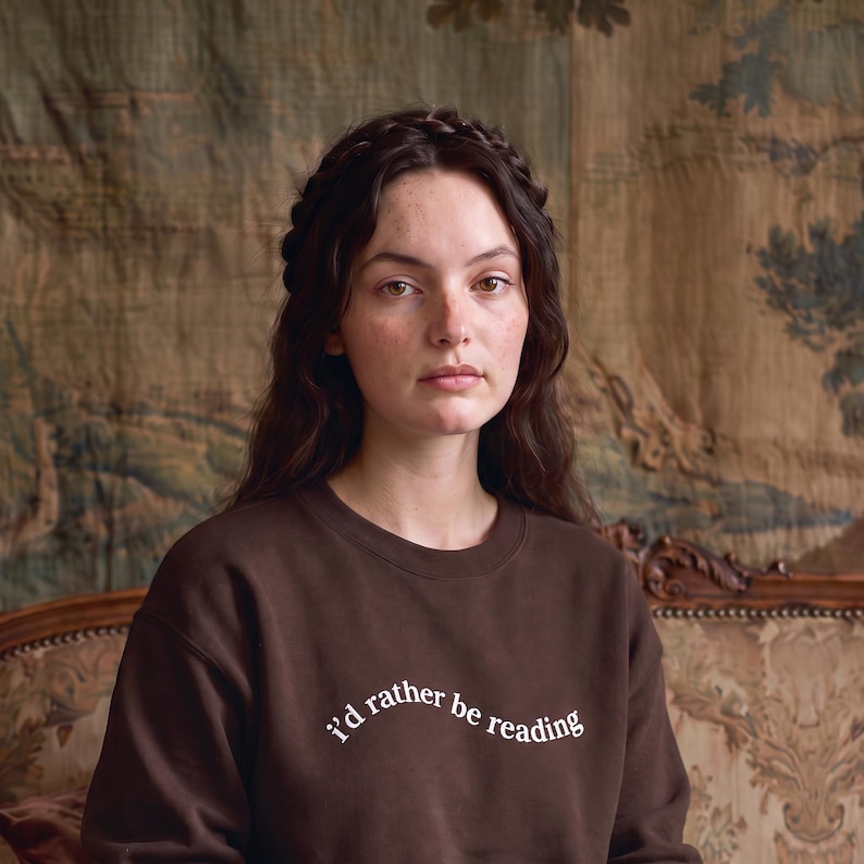 May include: A dark brown crewneck sweatshirt featuring the white text "i'd rather be reading" printed in a curved, lowercase font across the chest. The garment has long sleeves and a ribbed neckline, providing a simple and functional design.