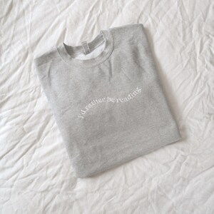 May include: A light gray crewneck sweatshirt featuring the white embroidered text "I'd rather be reading" arched across the chest. The garment has long sleeves, a ribbed collar, and a simple, clean design with a soft, heathered fabric texture.