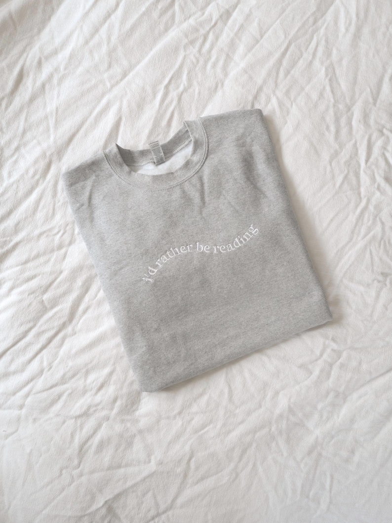 May include: A light gray crewneck sweatshirt featuring the white embroidered text "I'd rather be reading" arched across the chest. The garment has long sleeves, a ribbed collar, and a simple, clean design with a soft, heathered fabric texture.