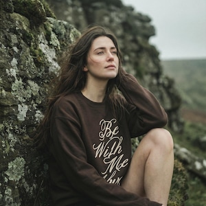 May include: A dark brown long-sleeve crewneck sweatshirt featuring the white cursive text "Be With Me" printed on the front. The garment has a ribbed neckline, cuffs, and hem, providing a standard fit with long sleeves and a classic pullover design.
