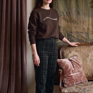 May include: A dark brown long-sleeve sweatshirt with the white text "i'd rather be reading" printed on the chest, paired with dark green and black plaid trousers and black flat shoes with a single strap across the top of the foot.