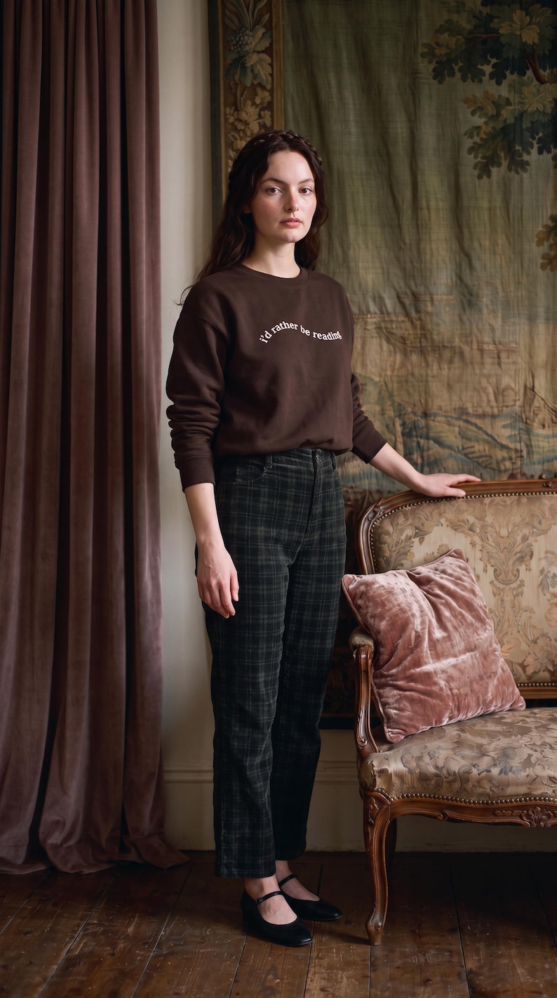 May include: A dark brown long-sleeve sweatshirt with the white text "i'd rather be reading" printed on the chest, paired with dark green and black plaid trousers and black flat shoes with a single strap across the top of the foot.