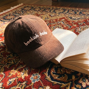 Puede incluir: Una gorra de béisbol de pana marrón con el texto blanco bordado "bookish babe" en el panel frontal. La gorra tiene una corona estructurada, una visera curva y ojales cosidos, colocada junto a un libro abierto con páginas blancas.