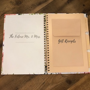 May include: A floral patterned wedding planner with a gold spiral binding. The planner has a pocket on the right side labelled "Gift Receipts". The front cover has the text "This book belongs to The Future Mr. & Mrs."