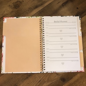 May include: A white and pink floral patterned bridal shower gift log book with a gold spiral binding. The book has a tabbed section for each gift and includes space to write the gift, from whom it came, and a thank you note.
