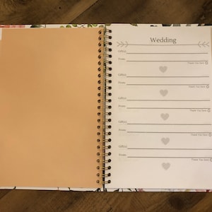 May include: A white spiral bound notebook with a floral design on the cover. The notebook has a section labelled "Wedding" with spaces to record gifts received, the giver's name, and a thank you note.