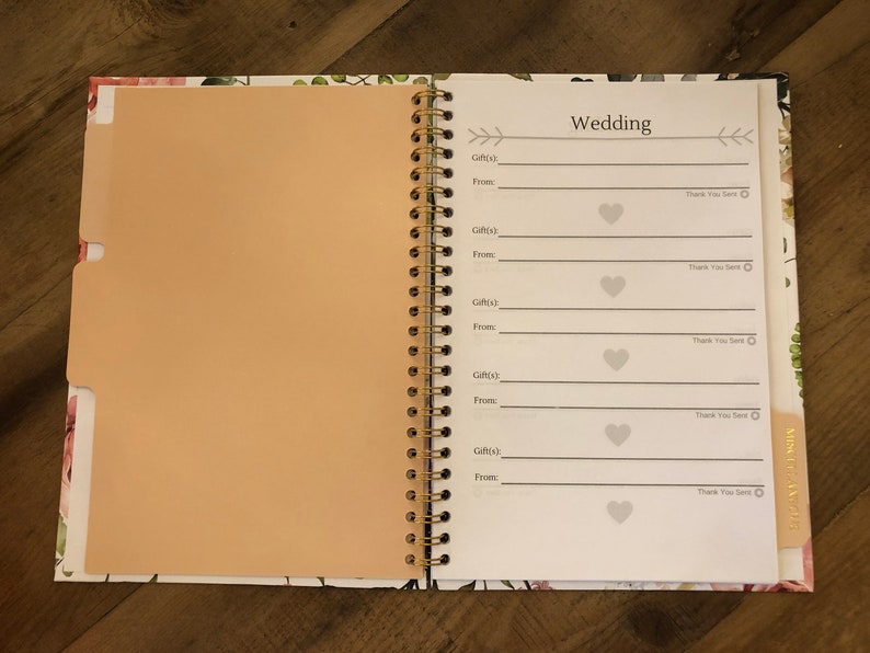 May include: A white spiral bound notebook with a floral design on the cover. The notebook has a section labelled "Wedding" with spaces to record gifts received, the giver's name, and a thank you note.