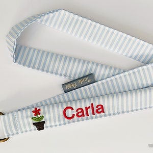 Op de afbeelding: Een blauw-wit gestreept koord met een metalen ring, een rode bloem en de naam "Carla" geborduurd in rood. Het koord heeft een klein label met de tekst "Tante Pauli".
