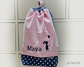 May include: Pink and blue drawstring bag with white polka dots and gingham pattern. The bag has a silhouette of a girl with a balloon and the name "Maya" embroidered on the front. The bag is labeled "von Tante Polli".